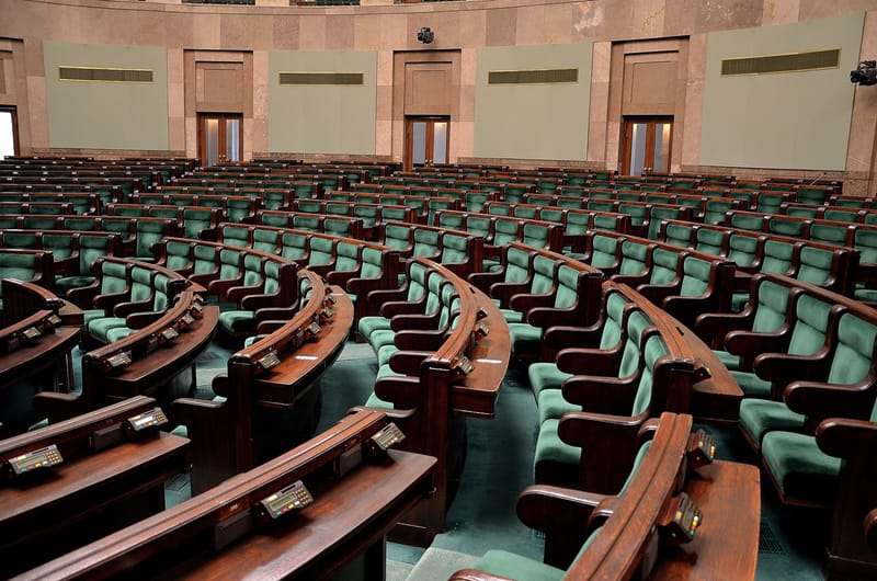 Pusta sala posiedzeń Sejmu: półkoliste rzędy drewnianych ław z zielonymi fotelami i pulpitami do głosowania.
