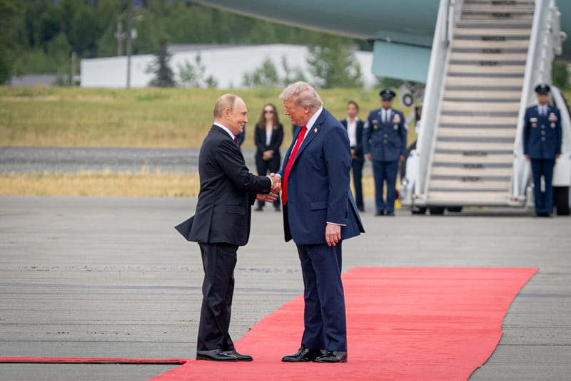 Donald Trump i Władimir Putin na lotnisku wojskowym w Alasce; obaj w ciemnych garniturach, w tle samoloty i flagi.